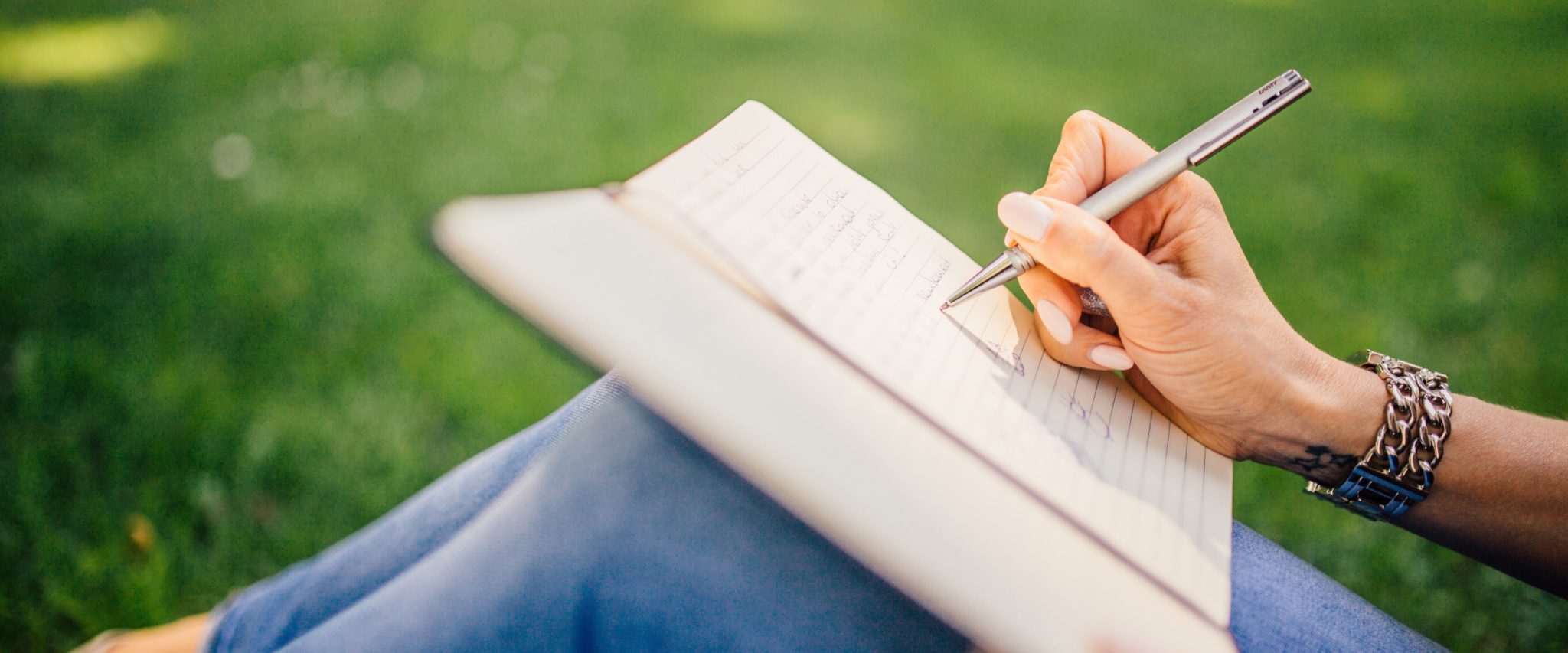 A conservation communicator writes in her notebook while sitting on green grass outdoors.