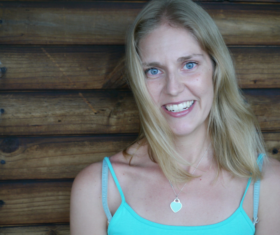 A woman with long blonde hair smiling, wearing a light blue tank top and a silver heart-shaped pendant necklace, standing against a wooden wall.