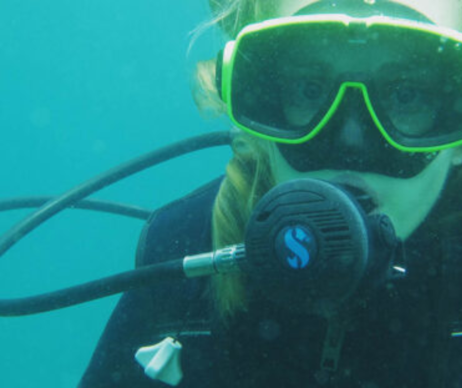 A scuba diver underwater, wearing neon green goggles and breathing apparatus, with braided hair visible inside the mask.
