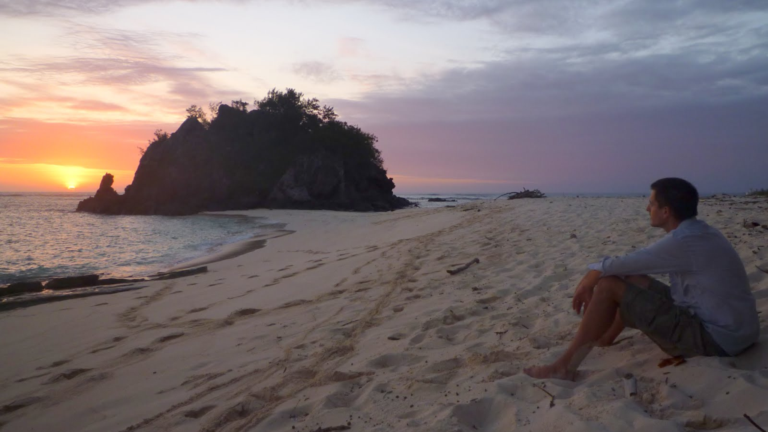 Dr Nick Askew sitting on a beach at sunset in Fiji, where he worked abroad in conservation for BirdLife International.