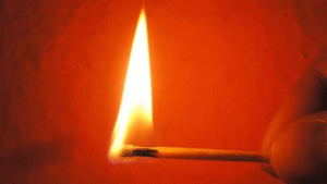 Close-up of a lit matchstick with a bright flame against a red-orange background.