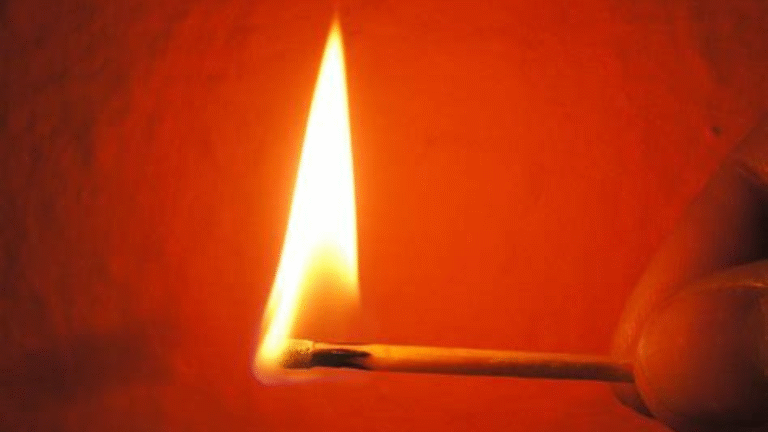 Close-up of a lit matchstick with a bright flame against a red-orange background.