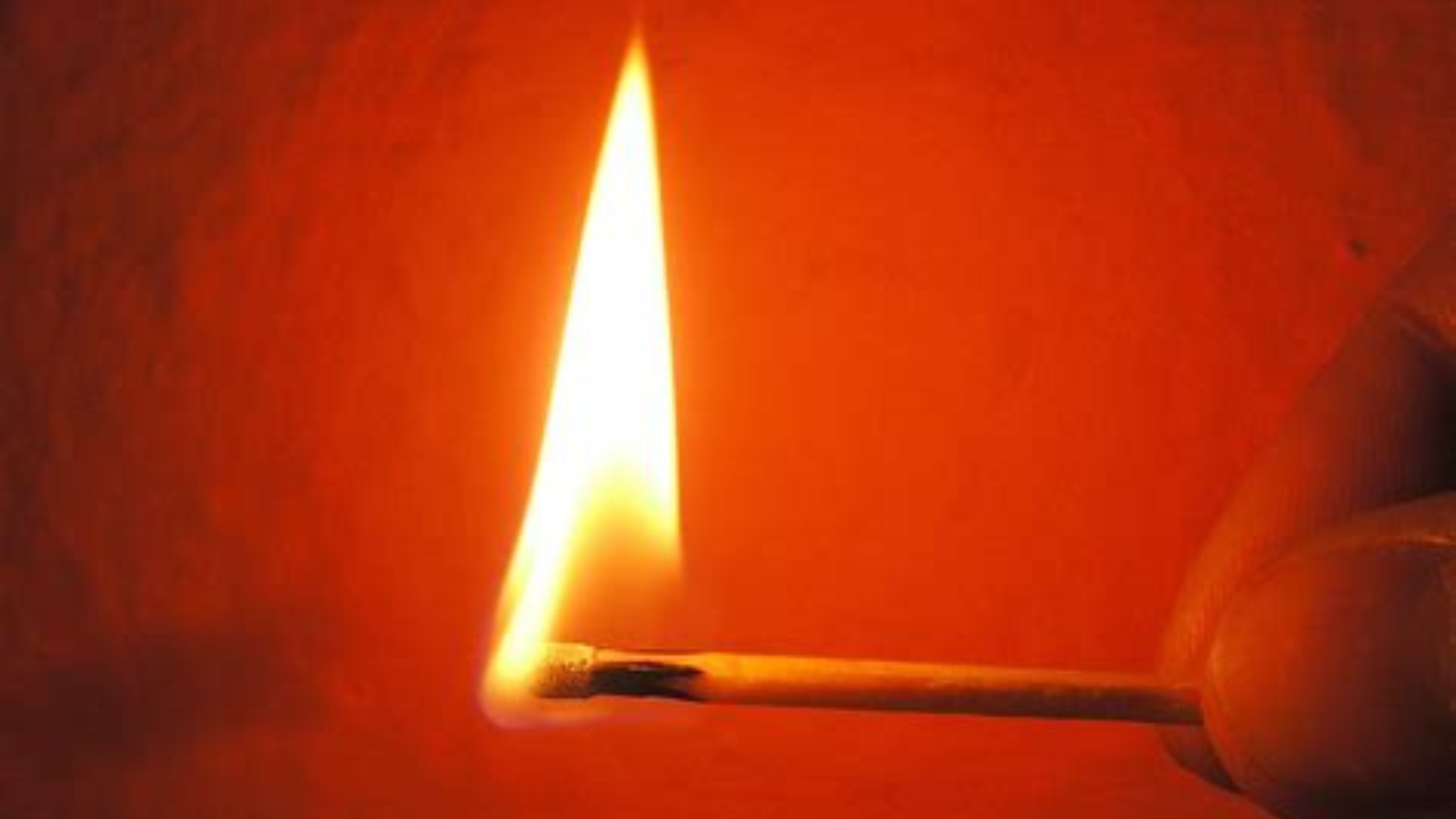 Close-up of a lit matchstick with a bright flame against a red-orange background.