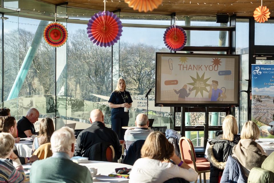 A woman stands presenting the work of the Vale Coastal Form to a room of stakeholders.