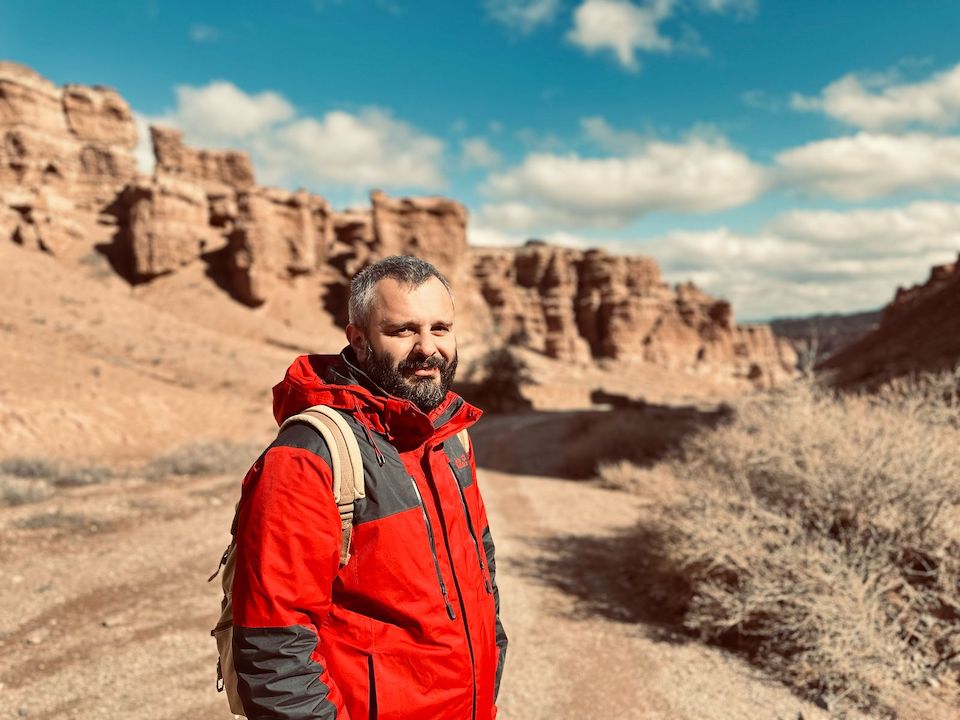 Man in a red jacket with a backpack standing on a path in a rocky canyon under a blue sky.