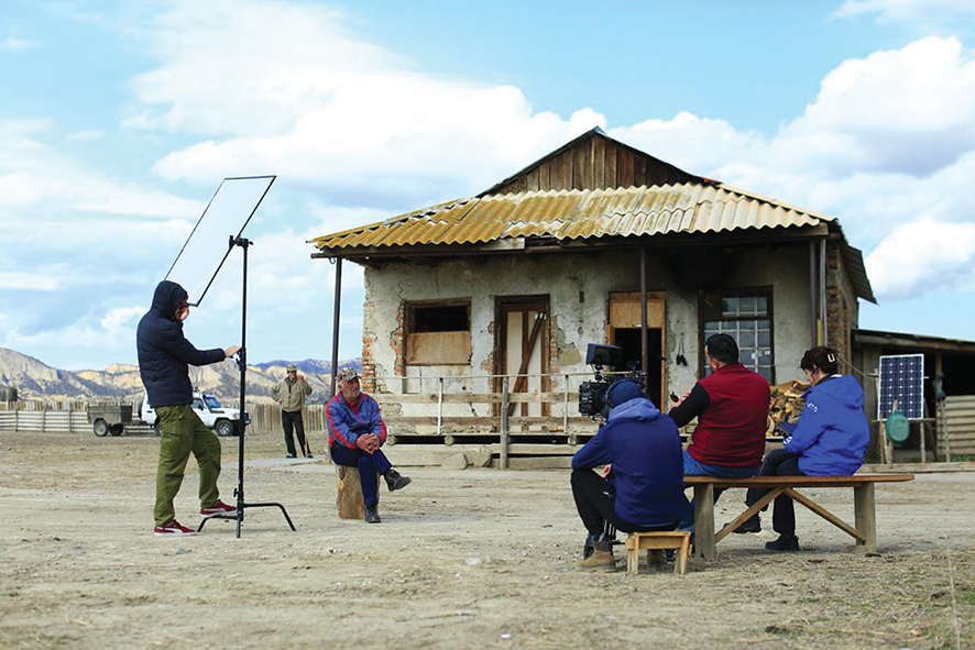 Film crew shooting an interview outdoors in front of a rustic, weathered house. 