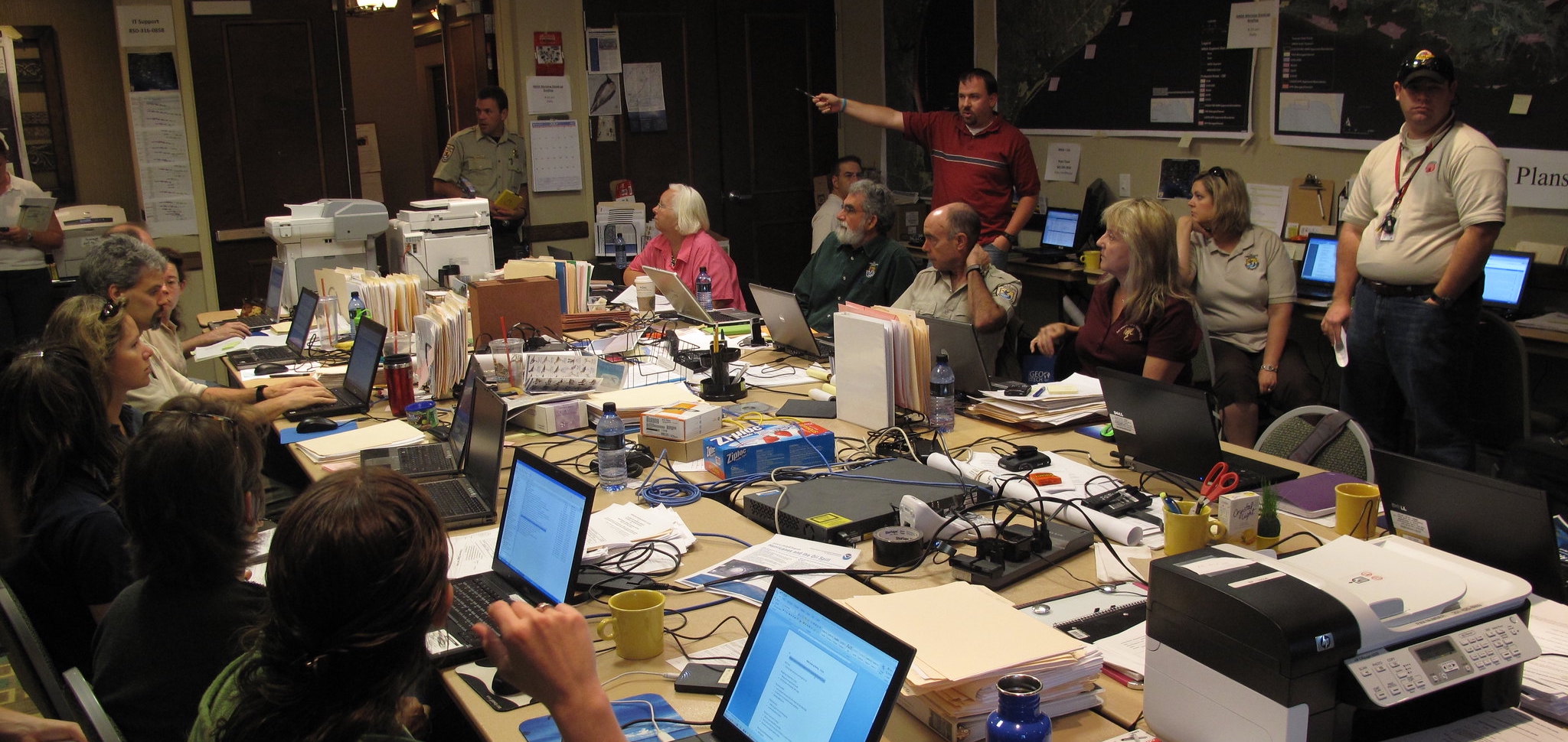 A GIS Analyst standing at a large table addressing a planning team who are seated at the table with laptops in front of them.