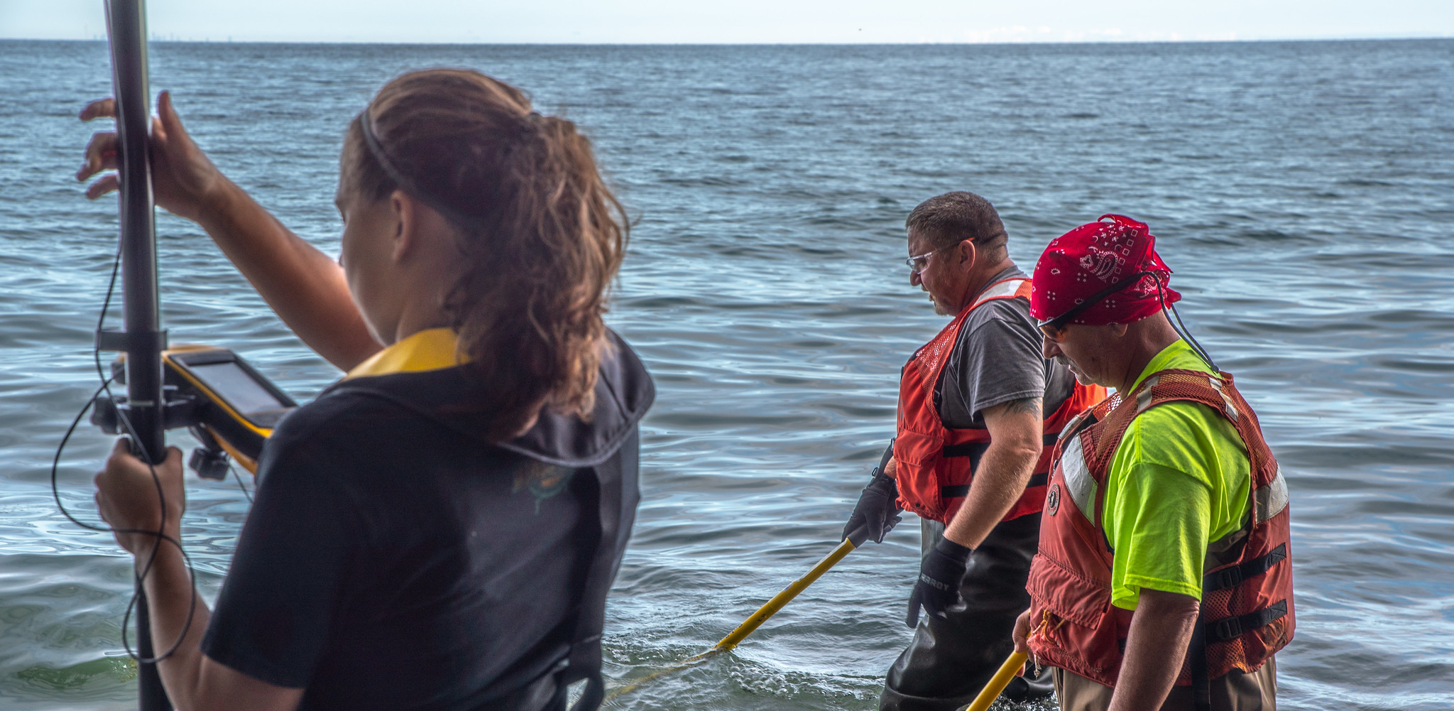 A woman and two men wearing safety vests standing in water and holding equipment. Many GIS Analyst duties are desk-based, but some GIS Analysts also support field data collection.