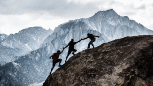 Three hikers climbing a steep rocky slope; one person at the top extends a hand to help the others, symbolising teamwork and mutual support in a challenging environment.