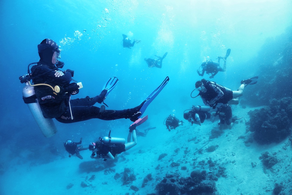 A group of scuba divers in full diving gear swim together underwater, surrounded by vibrant blue water with coral reefs visible on the seabed.