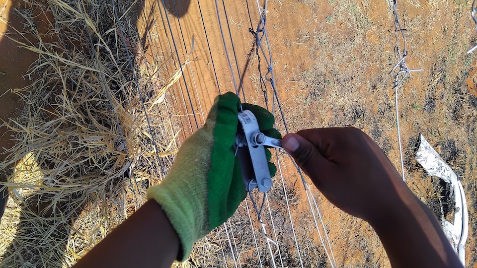 A person adjusts a boundary fence using a tensioning tool in a dry, grassy landscape.