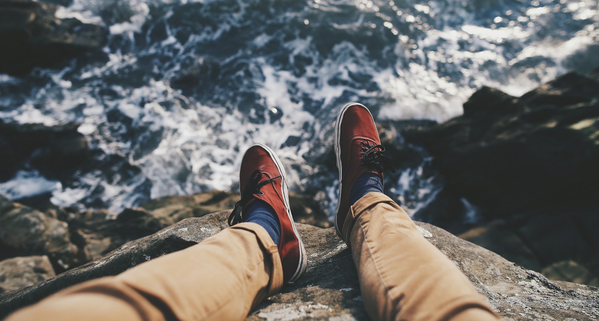 A pair of legs with trousers and shoes dangling over a cliff with the ocean below. Getting ready to work abroad in conservation can feel like jumping off a cliff into the unknown!