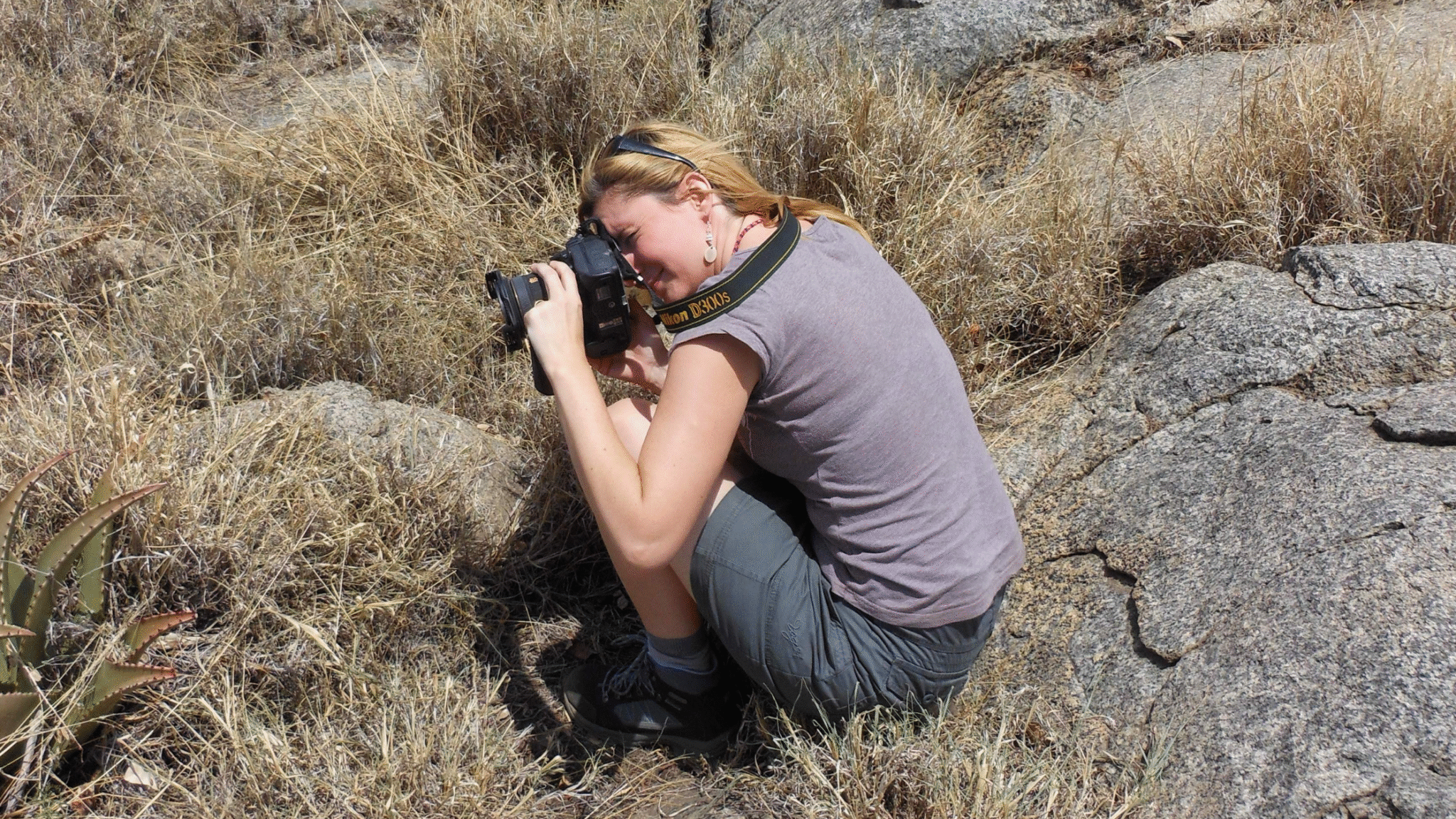 A woman crouched down in the grass holding a camera to her eye, photographing wildlife in the Serengeti.