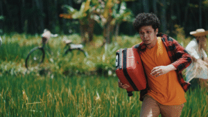 A man running anxiously through a green field while carrying a bright red suitcase. Photo by Rendy Novantino on Unsplash.