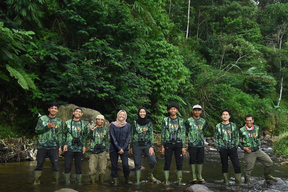 A group of nine people wearing wellies and outdoor attire, standing side by side in very shallow water in the forest, with trees in the background, all looking at the camera and smiling.