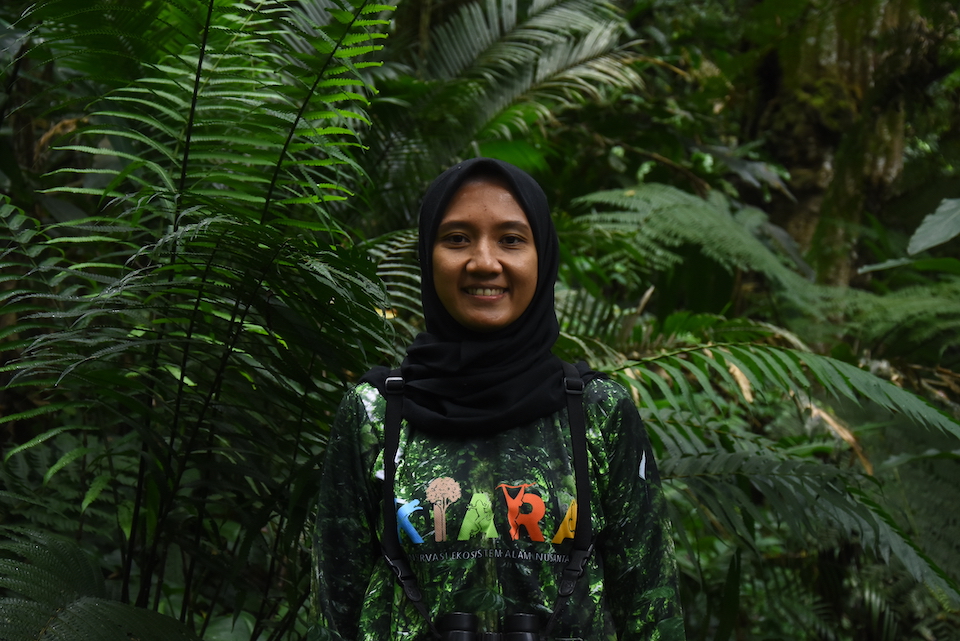 A woman, Rahayu Oktaviani, standing in front of plants, with binoculars around her neck, facing the camera and smiling.