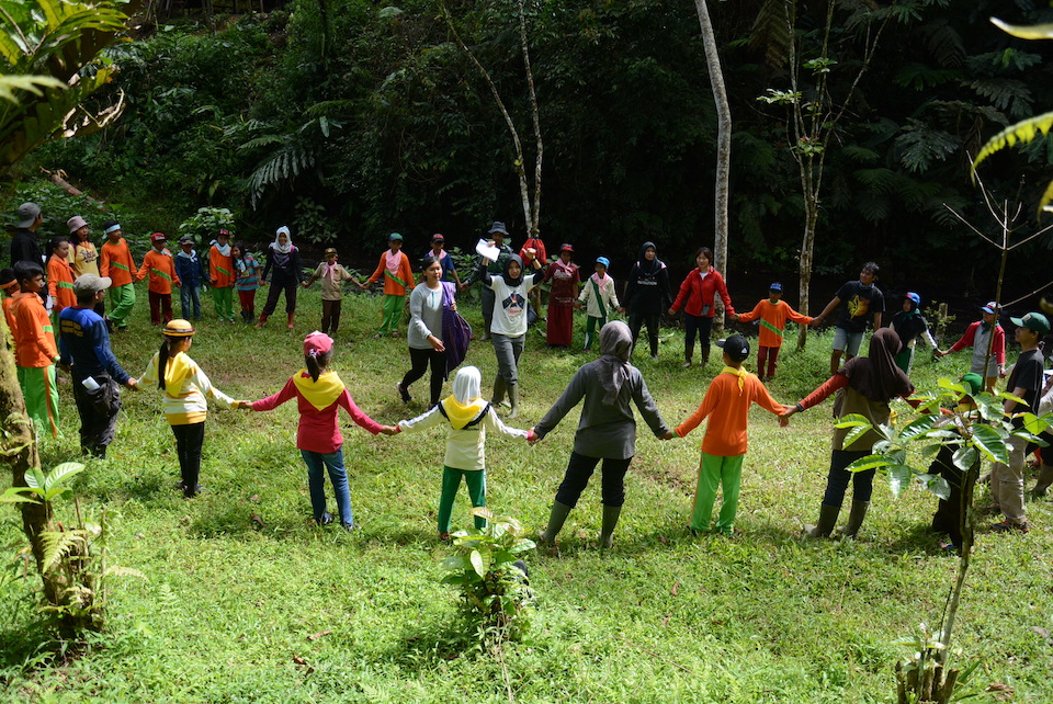 A large group of people stand together in a field, all holding hands in a circle, with two women in the middle of the circle.