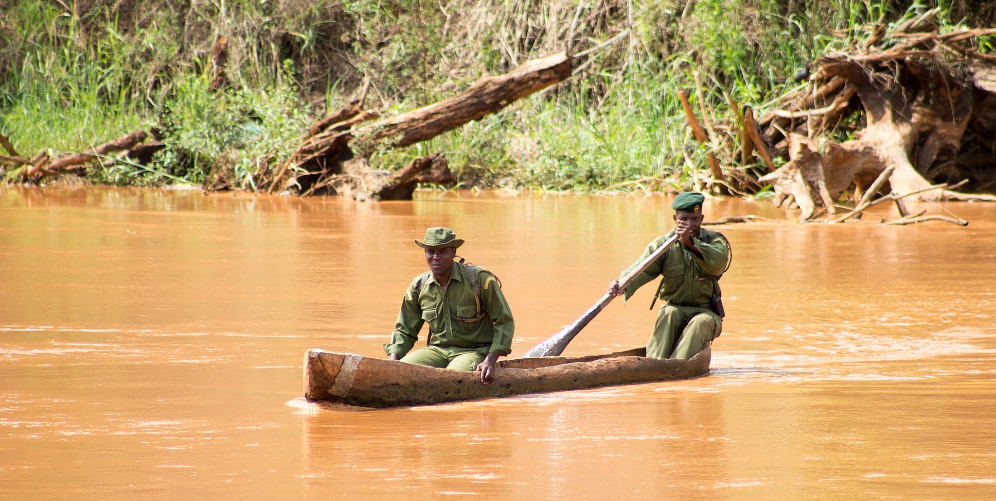 Two men in uniform paddling a dugout canoe across a brown river. Rangers are one example of a role within community-based conservation careers.
