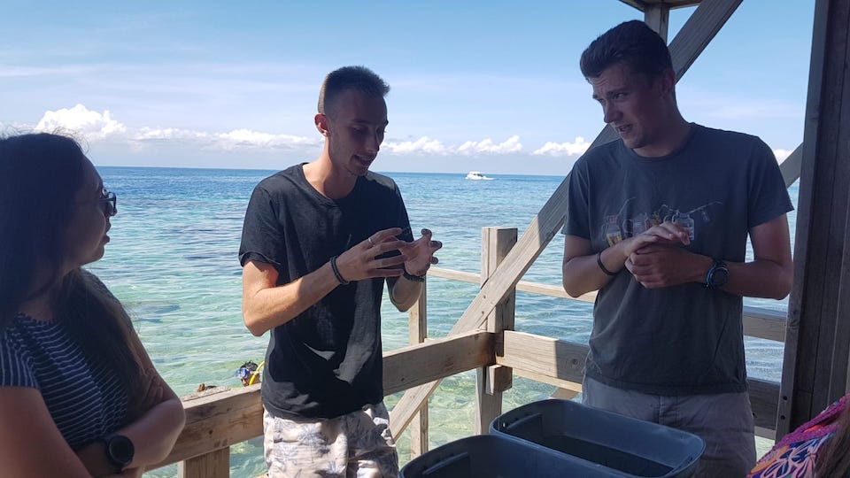 Three people standing on a wooden deck by the sea, engaged in conversation. A boat is visible in the distance on the clear blue water under a sunny sky.