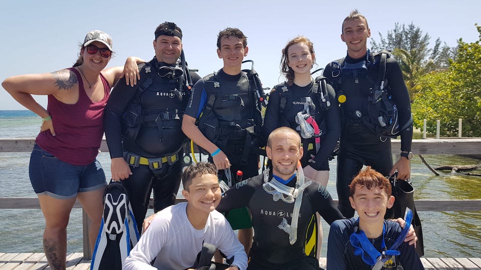 A group of seven people, some in scuba diving gear and some in casual clothing, pose together on a wooden dock near the water with trees in the background.